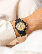 Black rubber strap with white contrast stitching on Omega x Swatch MoonSwatch Mission to the Moon 1969, worn on wrist, cream dial with gold-tone indices and pushers, black bioceramic case