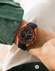 Black rubber strap with diagonal ribbed texture on Omega x Swatch MoonSwatch Mission on Earth Lava, wrist shot against cream fabric background, black dial with orange chronograph hands, dark green jacket with white button visible at left and lower right