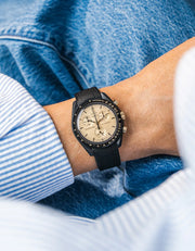 Plain black rubber strap on Omega x Swatch MoonSwatch Mission to the Moon 1969, wrist shot against denim jacket background, cream dial with silver-tone pushers, black bioceramic case