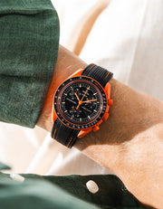 Black rubber strap with orange contrast stitching and diagonal ribbed texture on Omega x Swatch MoonSwatch Mission on Earth Lava, wrist shot against cream fabric background, black dial with orange chronograph hands, dark green jacket with white button visible at left and lower left
