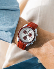 Burgundy rubber strap with diagonal ribbed texture on Omega x Swatch MoonSwatch Mission to Pluto, wrist shot against cream fabric background, white dial with burgundy subdials and burgundy tachymeter bezel, light grey bioceramic case, blue chambray shirt visible at left