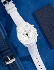 Flat lay of MoonSwatch Mission to the Moonphase with white vertech strap and accessories in background