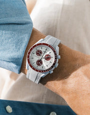 Grey rubber strap with cross-hatched ribbed texture on Omega x Swatch MoonSwatch Mission to Pluto, wrist shot against cream fabric background, white dial with burgundy subdials and burgundy tachymeter bezel, light grey bioceramic case, blue chambray shirt with white button visible at lower left