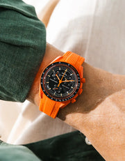 Lava orange rubber strap with vertical ribbed texture on Omega x Swatch MoonSwatch Mission on Earth Lava, wrist shot against cream fabric background, black dial with orange chronograph hands, dark green jacket visible at upper and lower left